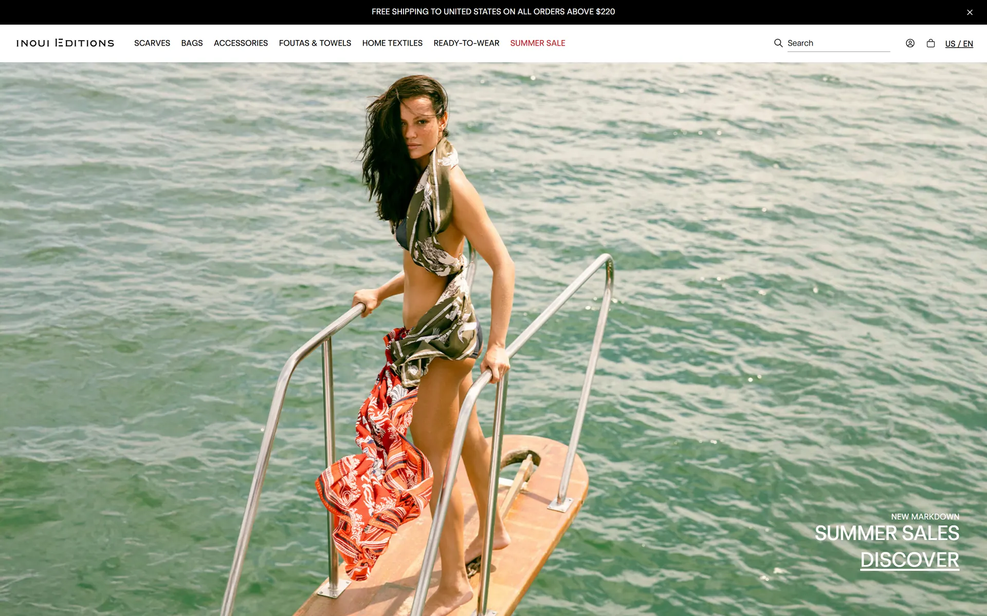 The Inoui Editions homepage on desktop: a full-bleed campaign image of a woman in a printed silk pareo on a boat over turquoise water, with the category navigation and a Summer Sales call to action